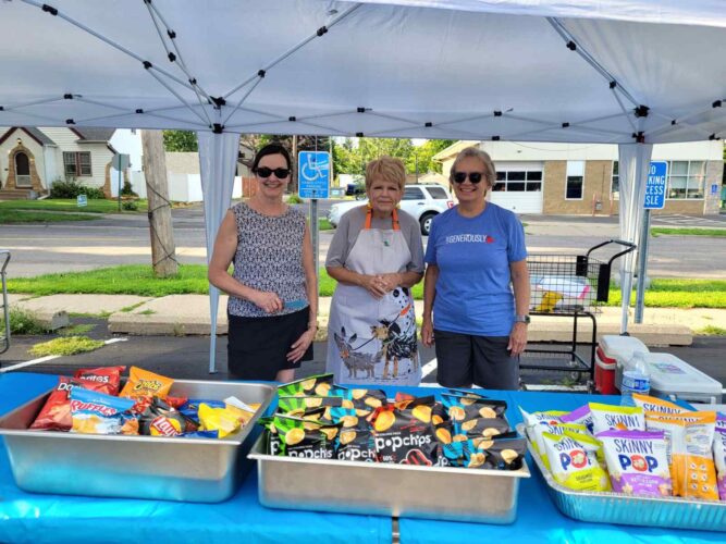 Three volunteers smile beneath a canopy behind a table filled with assorted bags of chips at an outdoor community event.