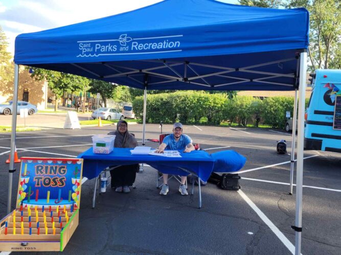 Two Saint Paul Parks and Recreation staff members welcome visitors beneath a blue canopy, with a colorful ring toss game set up nearby.